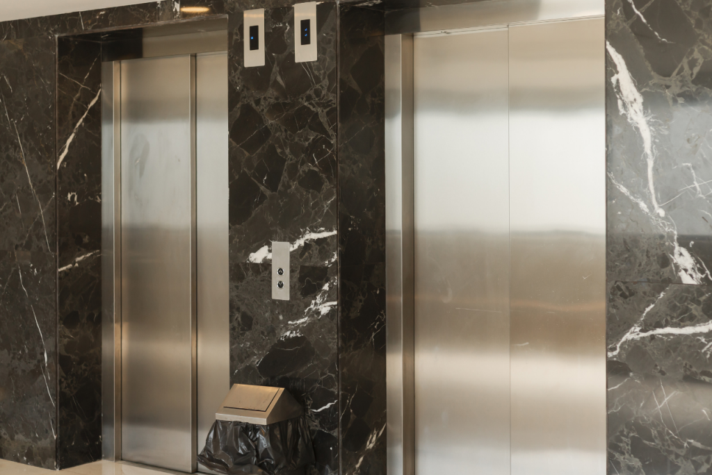 Zwei geschlossene Edelstahl-Aufzugstüren in einer eleganten Lobby mit dunkler Marmorwand und Anzeigeelementen darüber.