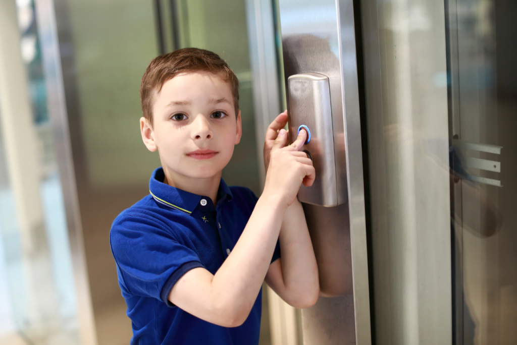 Junge drückt einen beleuchteten Aufzugknopf an einer modernen Edelstahl-Aufzugsanlage und blickt in die Kamera.