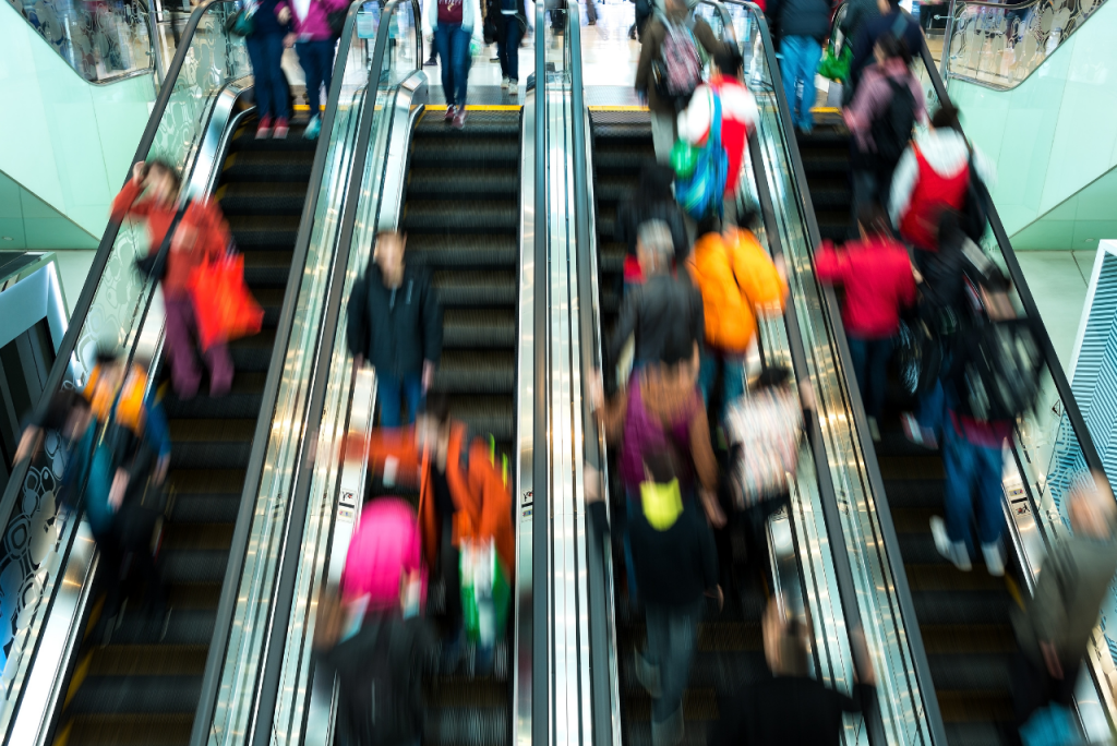 Viele Menschen bewegen sich auf parallelen Rolltreppen in einem belebten Einkaufszentrum, dargestellt mit deutlicher Bewegungsunschärfe.
