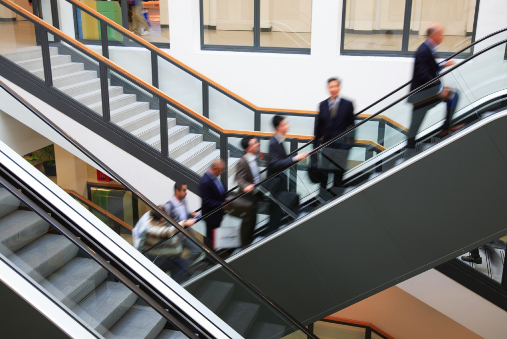 Mehrere Personen in Businesskleidung fahren eine Rolltreppe hinauf, aufgenommen mit leichter Bewegungsunschärfe in einem modernen Gebäude.